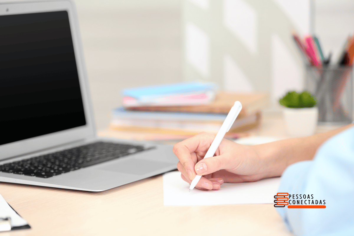 Mão segurando caneta, anotando ideias em caderno, com tela de notebook ao fundo exibindo um documento de copywriting